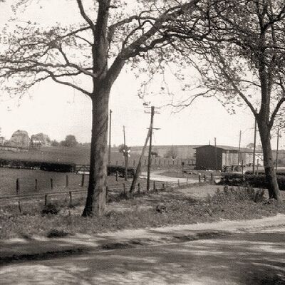 Das Foto von 1960 zeigt im Hintergrund den Bahnhof Schönkirchen vom Holzkatenweg aus gesehen in Blickrichtung Blomeweg. Das Gelände links und rechts vom Blomeweg ist noch unbebaut. Am Bahnhof erkennt man rechts das Bahnwärterhäuschen und links das Wartungs- und Materialhaus.